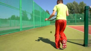 Genç bir çocuk tenis kortunun etrafında raketle koşup topa vuruyor..