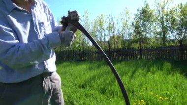 Mavi gömlekli ve gri iş pantolonlu bir adam güneşli bir yaz gününde tahta bir direkte tırpan keskinleştirir..