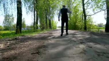 Pantolon, gömlek ve kazak giymiş, boynunda kulaklık olan yaşlı bir adam, huş ağacında asfalt bir yolda kaykay yapmayı öğreniyor..