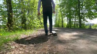 Pantolon, gömlek ve kazak giymiş, boynunda kulaklık olan yaşlı bir adam, huş ağacında asfalt bir yolda kaykay yapmayı öğreniyor..