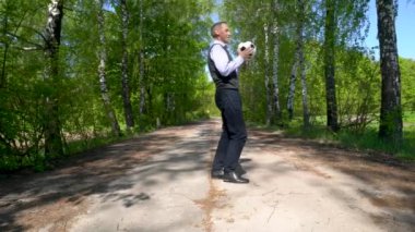 Mavi çizgili tişört ve süveter giyen yaşlı bir adam, siyah pantolon ve siyah botlar giymiş, huş ağacından bir sokakta diziyle futbol topunu tekmeliyor..