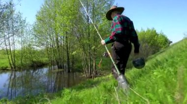 Erkek bir balıkçı yeşil çimlerle kaplı tepeden aşağı elinde olta kamışı ve kovayla göle doğru yürüyor. Kovboy şapkası, ekose gömlek, gri pantolon ve lastik çizme giyiyor. Dışarıda güneşli bir gün ve bir sürü sivrisinek var..