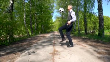 Mavi çizgili tişört ve süveter giyen yaşlı bir adam, siyah pantolon ve siyah botlar giymiş, huş ağacından bir sokakta diziyle futbol topunu tekmeliyor..