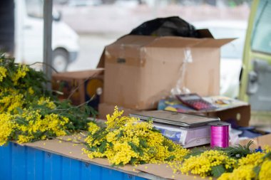 Pullu bir masa ve satılık bir kutu mimoza çiçeği. Çiçekler sarı ve masada başka eşyalar da var..