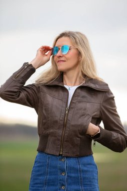 Kahverengi deri ceket ve kot pantolon giyen bir kadın gülümsüyor ve güneş gözlüğü takıyor. Güven ve tarz kavramı