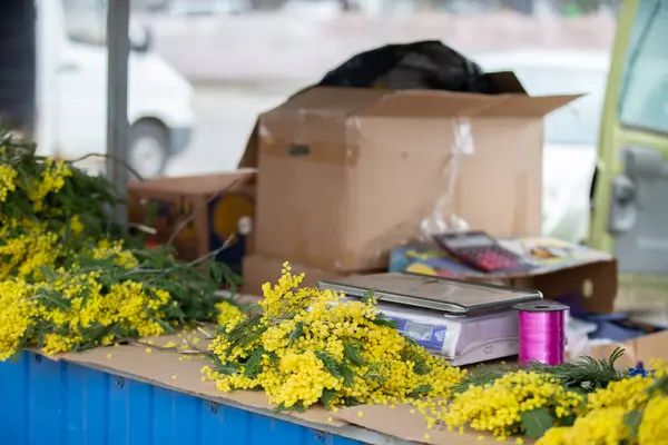 Pullu bir masa ve satılık bir kutu mimoza çiçeği. Çiçekler sarı ve masada başka eşyalar da var..