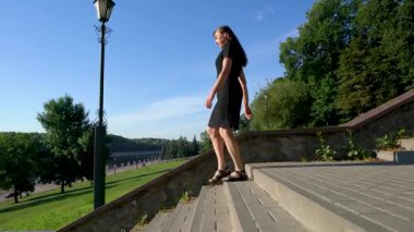 Siyah elbiseli yaşlı bir kadın merdivenlerden inip park alanındaki sete doğru yürür..