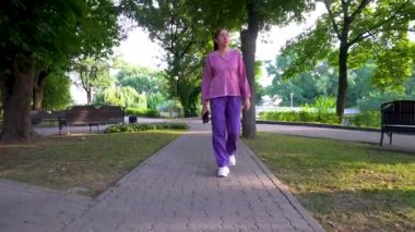 Günlük giysiler içinde yetişkin bir kadın park alanında yürüyor. Elinde bir telefon tutuyor..