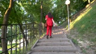 Kırmızı elbiseli bir kadın şehir parkının merdivenlerinden çıkıyor. Arkadan görüntüle.