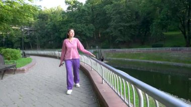 Güneşli bir günde güzel bir parkta günlük giysiler giymiş yetişkin bir kadın yürüyor. Keyfi yerinde..