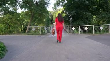 Kırmızı elbiseli uzun boylu bir kadın şehir parkında ilerler. Arkadan görüntüle.