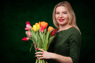 Orta yaşlı güzel bir sarışın kadın elinde bir buket lale çiçeği tutuyor, gülümsüyor ve karanlık bir arka planda kameraya bakıyor..