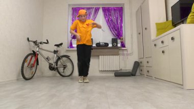 Genç bir çocuk mor süslemelerle süslenmiş rahat bir odada dans ediyor. Hareket becerilerini ve ritmini sergileyen bir bisiklet duvara yaslanır..