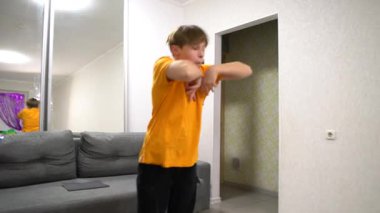 Modern bir oturma odasında, turuncu gömlekli genç bir çocuk dans yeteneklerini sergiliyor. Kendine güvenerek hareket eder, bir aynanın karşısında rutinini çalışırken anın tadını çıkarır..