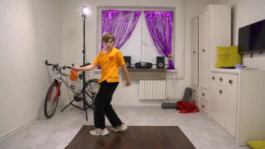 Canlı bir iç mekanda, genç bir dansçı renkli dekorasyonlar ve bir bisikletle çevriliyken etkileyici hareketler sergiler. Parlak perdeler ve minimalist mobilyalar yaratıcı bir atmosfer yaratır..