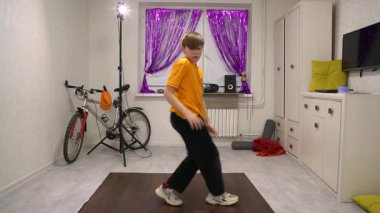 Genç bir çocuk dans figürlerini parlak perdelerle süslenmiş renkli bir odada sergiliyor. Koreografisini prova ederken müzik hafifçe çalıyor, odaklanmış ve enerjik..