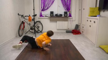 Parlak turuncu gömlekli bir çocuk, rahat bir oturma odasında, ahşap bir zeminde break dans yapıyor. Mekanda bir bisiklet, hoparlörler ve mor perdeler var..