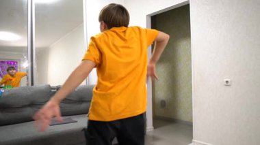 Turuncu tişörtlü genç bir çocuk, dansını açık renkli duvarları ve rahat bir kanepesi olan parlak bir oturma odasında sergiliyor. Ayna onun hareketlerini yansıtıyor..