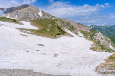 Çiçekli çayırlar ve karla kaplı dağlık tepeleri olan kayan tepelerin manzaralı panoramik manzarası. Güzel güneşli bir gün mavi gökyüzü ve baharda bulutlar.