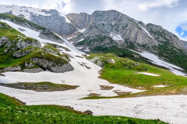 Çiçekli çayırlar ve karla kaplı dağlık tepeleri olan kayan tepelerin manzaralı panoramik manzarası. Güzel güneşli bir gün mavi gökyüzü ve baharda bulutlar.