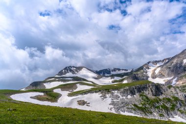 Çiçekli çayırlar ve karla kaplı dağlık tepeleri olan kayan tepelerin manzaralı panoramik manzarası. Güzel güneşli bir gün mavi gökyüzü ve baharda bulutlar.