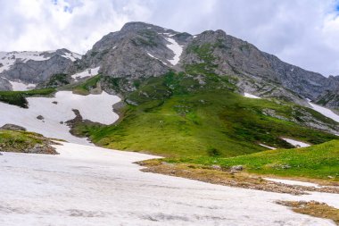 Çiçekli çayırlar ve karla kaplı dağlık tepeleri olan kayan tepelerin manzaralı panoramik manzarası. Güzel güneşli bir gün mavi gökyüzü ve baharda bulutlar.