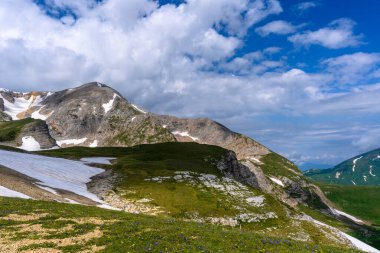 Çiçekli çayırlar ve karla kaplı dağlık tepeleri olan kayan tepelerin manzaralı panoramik manzarası. Güzel güneşli bir gün mavi gökyüzü ve baharda bulutlar.
