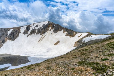 Çiçekli çayırlar ve karla kaplı dağlık tepeleri olan kayan tepelerin manzaralı panoramik manzarası. Güzel güneşli bir gün mavi gökyüzü ve baharda bulutlar.