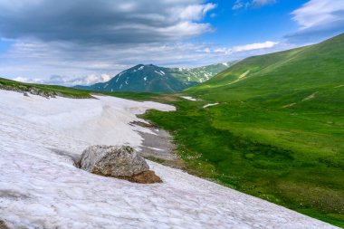 Çiçekli çayırlar ve karla kaplı dağlık tepeleri olan kayan tepelerin manzaralı panoramik manzarası. Güzel güneşli bir gün mavi gökyüzü ve baharda bulutlar.
