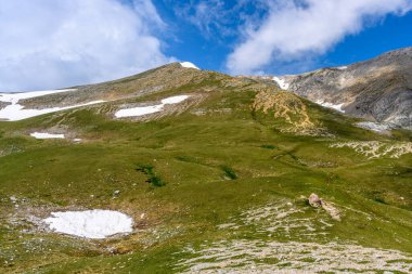 Çiçekli çayırlar ve karla kaplı dağlık tepeleri olan kayan tepelerin manzaralı panoramik manzarası. Güzel güneşli bir gün mavi gökyüzü ve baharda bulutlar.