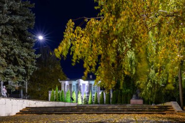 Ulyanovsk, Rusya - 20 Ekim 2024: Sonbahar yaprakları dökülen bir şehir parkında sakin bir gece sahnesi. Arka plandaki tarihi bina huzurlu şehir atmosferine cazibe katıyor..