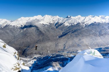 Bir adam uçurumun kenarındaki teleferiğin üzerinde yürüyor. Aşırı bir cazibe. Resmedilmeye değer karlı bir sıradağ heybetli bir şekilde duruyor açık mavi gökyüzüne karşı.