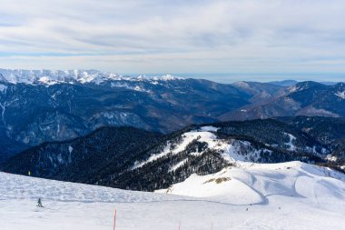 Bir dağ yamacında kayakçılar batıyor. Arka planda güzel karlı dağlar ve vadi manzarası var..