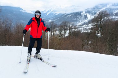 Bir kayakçı güvenli bir şekilde karla kaplı bir yamaçta poz verir ve dinamik bir kış manzarası yaratır. Karla kaplı dağlar ve ağaçlar görüntüye enerji katıyor.
