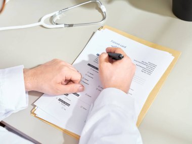Beyaz önlüklü, kimliği belirsiz bir erkek doktor klinikte tıbbi belgeler dolduruyor.