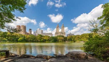 New York 'un arka planında gökdelenleri olan Central Park gölü beyaz bulutlu mavi gökyüzünün altında, ABD