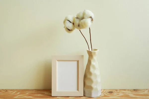 Square wooden picture frame and vase of soft cotton branch on wooden table. ivory wall background. blank message card, memo space, copy space