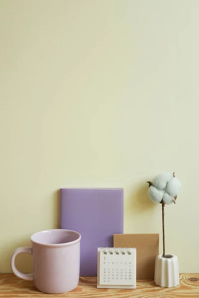 Purple diary notebook, mug cup, calendar, vase of cotton plant on wooden desk. ivory wall background. plan, workspace, office supply