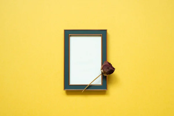 Empty navy photo frame and dry rose flower on yellow background. top view, copy space, minimalism