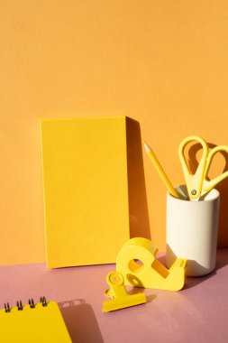 Ofis malzemeleri kırtasiyesi. Sarı defter, renkli kalem, makas, bant kesicisi, pembe masadaki şarjör. turuncu duvar arka planı. çalışma alanı
