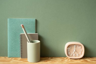 Kırtasiye malzemeleri. Defter, kalem tutacağı, tahta masadaki saat. Yeşil duvar arkaplanı. çalışma alanı
