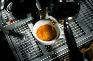 Bir espresso makinesinin damlama tepsisinin üzerinde duran beyaz kahve fincanındaki güzel altın kremayla yakın plan bir espresso. Görüntü, kahve hazırlama anını mükemmel bir şekilde yakalıyor..