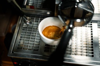 Bir espresso makinesinin damlama tepsisinin üzerinde duran beyaz kahve fincanındaki güzel altın kremayla yakın plan bir espresso. Görüntü, kahve hazırlama anını mükemmel bir şekilde yakalıyor..