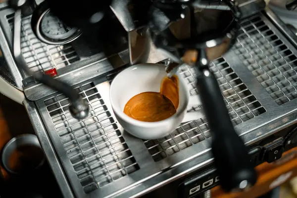 Bir espresso makinesinin damlama tepsisinin üzerinde duran beyaz kahve fincanındaki güzel altın kremayla yakın plan bir espresso. Görüntü, kahve hazırlama anını mükemmel bir şekilde yakalıyor..