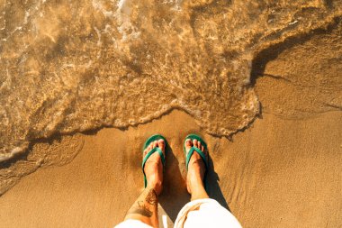 Yeşil parmak arası terlikler altın kumda dinleniyor. Plajda sakin bir yaz yürüyüşü olduğunu gösteren ayak izleri var.