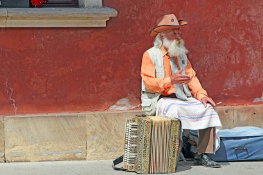 Varşova - Polonya. 27 Temmuz 2019: Akordeonun yanında oturan gri saçlı yaşlı adam. Akordiyon sigaralı yaşlı adamın yanında duruyor. Yaşlı müzisyen hayatını akordeon çalarak kazanıyor. Yaşlı sokak müzisyeni sigara içiyor.