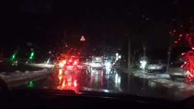 Yağmurlu bir gecede şehirde araba gezintisi. Arabanın ön camına yağmur yağdı. Gece şehir ışıkları. Akşam şehri boyunca at sürüyorum. Gece Araba Penceresi. Arka planda Gece Şehri Trafiği. Araba silecekleri yağmurda çalışıyor.