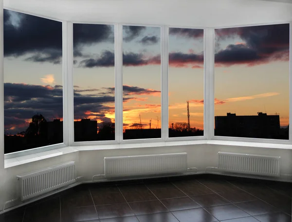 Panoramic window with view of red sunset above city buildings. Evening cityscape. Beautiful view from room window. Panoramic window overlooking the evening city. Twilight in city. Raising sun