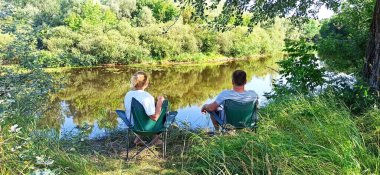 Evli çift nehir kenarında doğada dinleniyor. Karı koca nehirde vakit geçirir. Yaz tatili. Doğada yaz rahatlığı. Kadın ve erkek nehir kenarındaki sandalyelerde oturarak vakit geçiriyorlar.