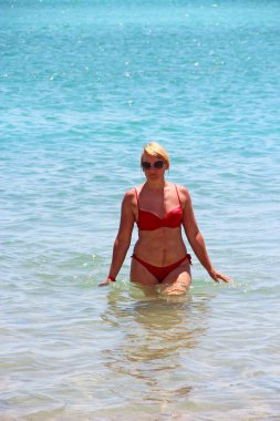 Denizde duran kırmızı mayolu kız. Kırmızı, modern mayolu, deniz kenarında dinlenen güzel bir kadın. 40 yaşındaki bir kadın yaz tatilinde ılık denizin tadını çıkarıyor.